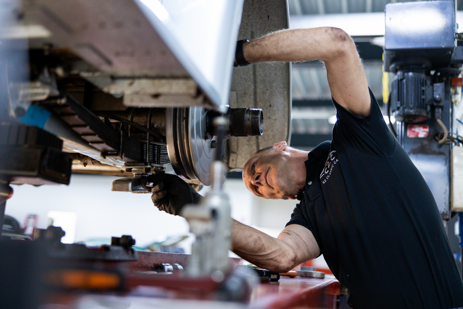 Jurgen inspiziert die Bremsen eines Oldtimers. Das Auto befindet sich auf einer roten Autohebebühne, um den Zugang zu erleichtern. Er führt Reparaturen an dem Oldtimer durch. Restaurierung ist einer der Dienste, die wir bei S2 Classic Cars anbieten, neben Coachbuilding, Dynamometer, Transport und Detailpflege