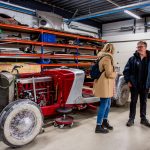 A pre war classic car with many parts stripped off. Still on its wheels. with two people standing to the right of it having a conversation